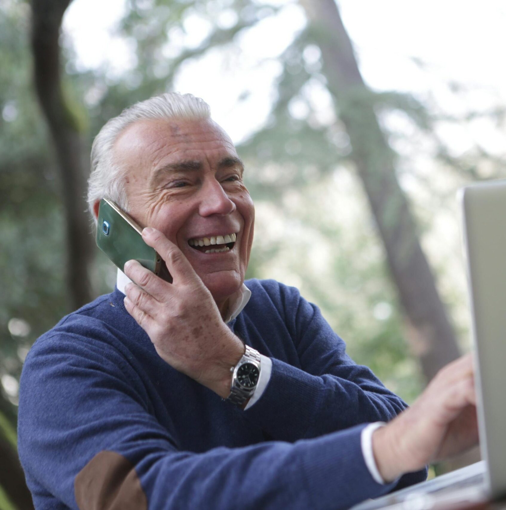 Happy senior man talking on phone and using laptop outdoors.