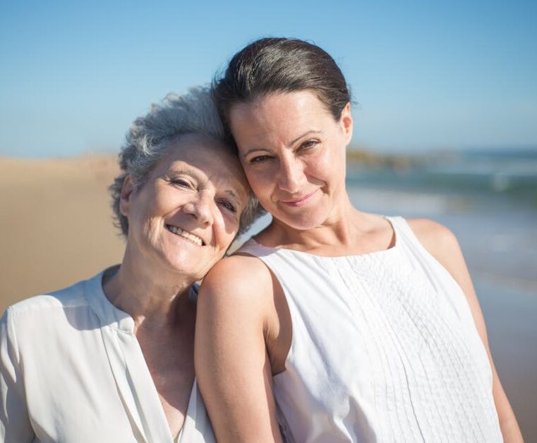 A senior mother and her adult daughter smiling together on a sunny beach in Portugal.